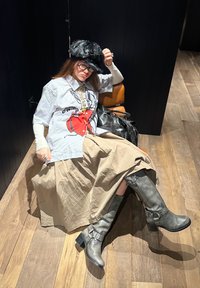 Valkoinen lyhythihainen paita, jossa on punainen graafinen printti, beige laskoshelma, ja harmaat polvipituiset saappaat, joissa on soljet. Istuu puulattialla.