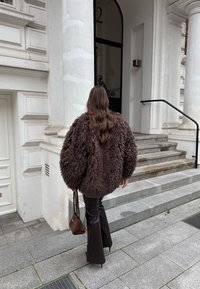 Jachetă maro din blană artificială, cu un design voluminos și texturat, asortată cu pantaloni negri eleganți și o geantă mică maro, urcând trepte din piatră.