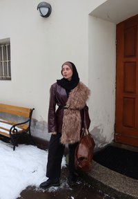 Femme portant une veste en cuir marron, un col en fourrure, un hijab noir et des bottes, tenant un sac marron, debout près d'une porte en bois et d'un banc couvert de neige.