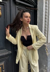 Κίτρινο σακάκι με ρίγες πάνω από μαύρη εφαρμοστή μπλούζα, συνδυασμένο με ταιριαστά παντελόνια. Τα αξεσουάρ περιλαμβάνουν ένα κολιέ σε σχήμα αστεριού.