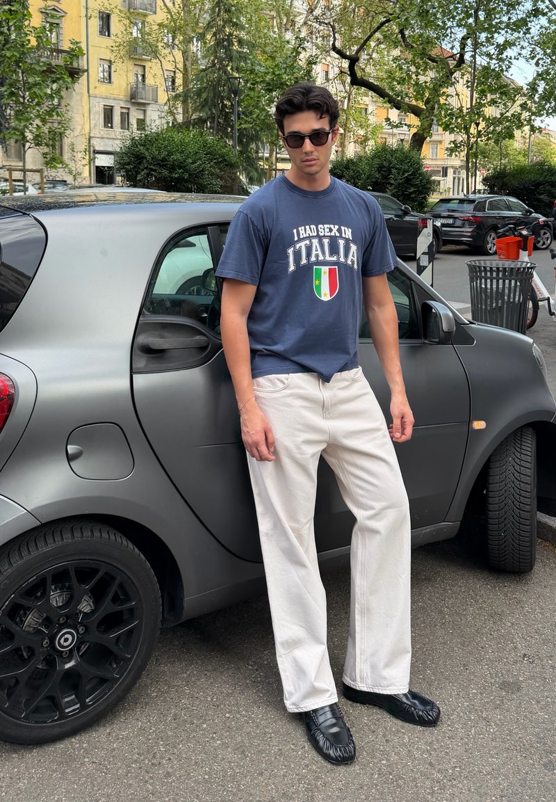 Jeune homme portant des lunettes de soleil, un t-shirt graphique bleu marine, un pantalon blanc et des chaussures noires, se tenant à côté d'une petite voiture grise dans une rue en ville.