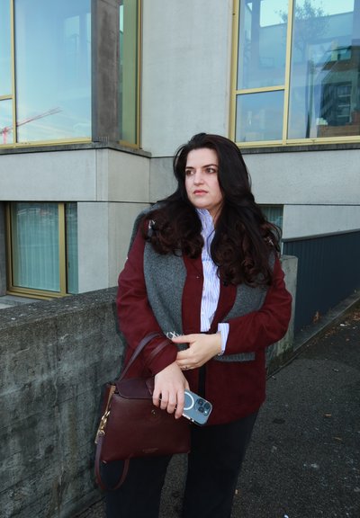 Chaqueta burdeos superpuesta sobre una bufanda gris, camisa blanca con rayas, sosteniendo un bolso granate y un teléfono inteligente. Cabello rizado oscuro, entorno al aire libre.