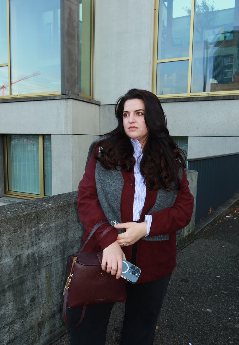 Veste bordeaux superposée à une écharpe grise, chemise blanche à rayures, tenant un sac à main marron foncé et un smartphone. Cheveux bouclés et foncés, cadre extérieur.