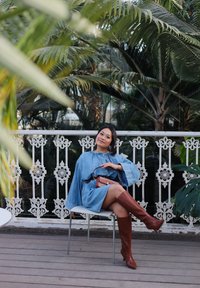 Robe en denim à manches larges, bottes marron jusqu'aux genoux et petit sac à main. Chaise contre une balustrade blanche décorative avec des plantes en fond.