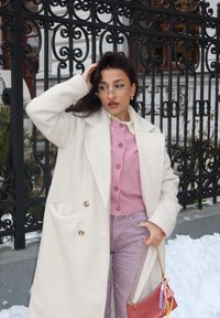 Γυναίκα με λευκή ζακέτα και ροζ πουλόβερ, κρατώντας μια μικρή κόκκινη τσάντα, στέκεται κοντά σε μια μαύρη περίφραξη από σφυρήλατο σίδερο με χιόνι στο έδαφος.