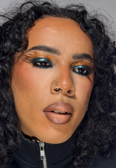 Primer plano de una persona con pelo rizado, maquillaje de ojos azul oscuro y naranja, pintalabios beige, un aro en la nariz y una camiseta negra con cremallera.