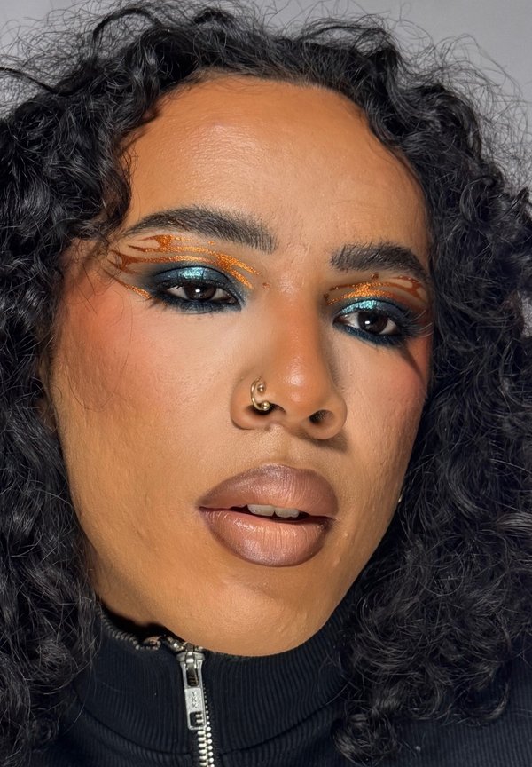 Primer plano de una persona con pelo rizado, maquillaje de ojos azul oscuro y naranja, pintalabios beige, un aro en la nariz y una camiseta negra con cremallera.