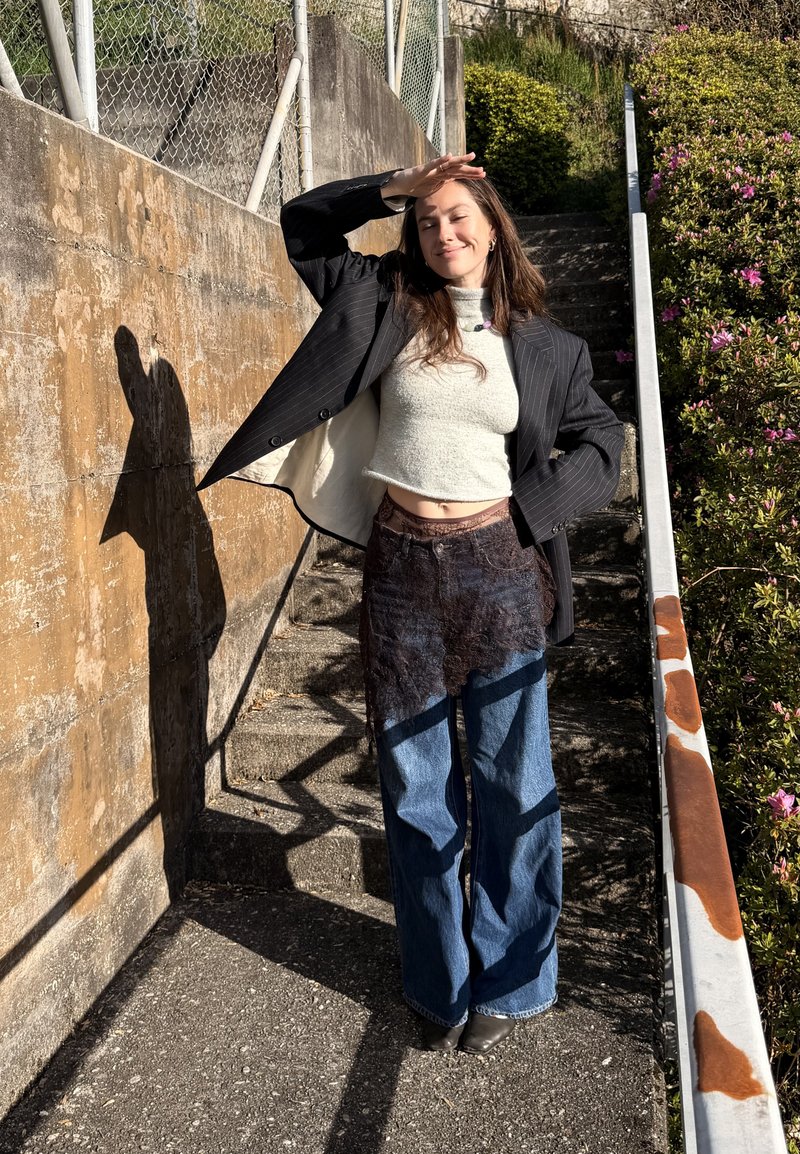 Jeune femme debout sur des marches extérieures, protège ses yeux du soleil, porte un blazer noir, un pull court, un pantalon avec plusieurs couches, et sourit les yeux fermés.