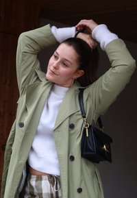 Ανοιχτό πράσινο trench coat με δομημένο σχέδιο, μαύρη τσάντα χειρός με χρυσές λεπτομέρειες, λευκή πλεκτή μπλούζα και καρό παντελόνι.