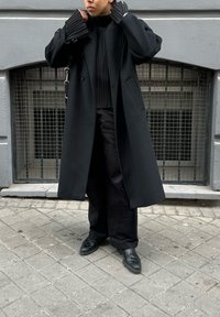 Μαύρο παλτό από μαλλί με υπερμεγέθη σχεδίαση, δομημένους ώμους και δύο μπροστινές τσέπες. Φορεμένο πάνω από ένα πλισέ μαύρο τοπ και φαρδιά παντελόνια.