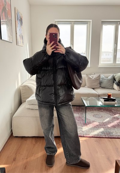 Chaqueta negra tipo puffer con acabado brillante, cierre frontal y mangas anchas, combinada con jeans gris de pierna ancha y zapatos marrones tipo slip-on.