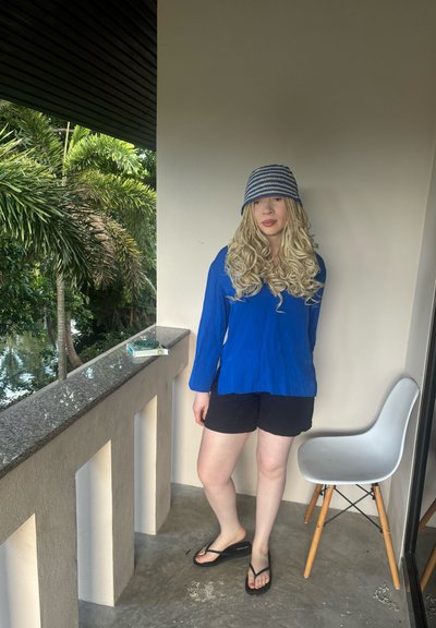 Modelo con una camisa de manga larga azul, pantalones cortos negros y chanclas, con un sombrero a rayas. Presenta cabello rubio ondulado, de pie junto a una barandilla de granito.