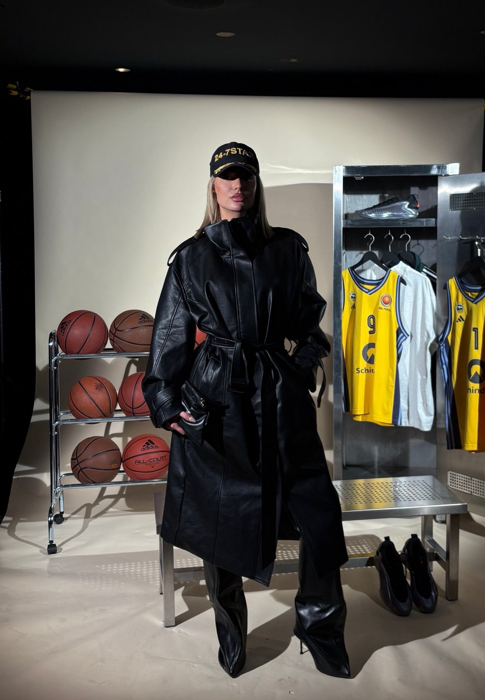 Trench coat de couro preto com cintura cingida, usado com calças de couro combinando e botas de salto. Um boné preto com letras amarelas está incluído.
