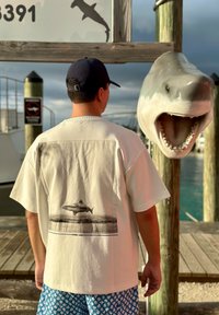 Homme portant un t-shirt et une casquette à thème requin, debout près d'un quai avec une grande sculpture de requin fixée à un poteau en bois.