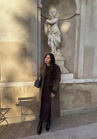 Long manteau marron sur un pull sombre, sac à main noir et bottines noires. Derrière, un mur en pierre avec une statue classique. Couleurs douces et atténuées.