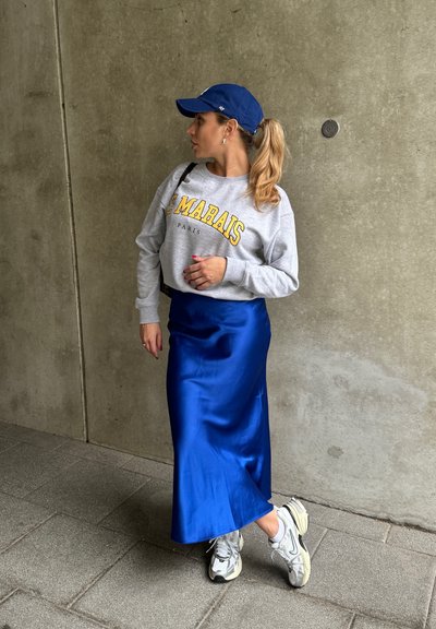 Sudadera gris con letras amarillas, falda de satén azul y zapatillas blancas con detalles grises. La persona está de pie contra una pared de concreto.