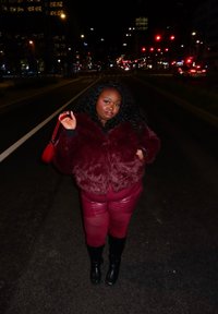 Γυναίκα με μπορντό γούνινο μπουφάν, δερμάτινο παντελόνι και μαύρες μπότες, στέκεται σε μια αστική οδό τη νύχτα, κρατώντας μια κόκκινη τσάντα ταιριαστή.