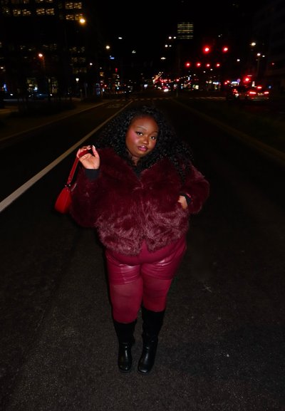 Mujer con chaqueta de piel color burdeos, pantalones de cuero y botas negras, de pie en una calle de la ciudad por la noche, sosteniendo un bolso rojo a juego.