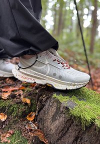 Osoba w białych butach do biegania Nike Trail stąpa po pokrytym mchem pniu drzewa w lesie, wśród opadłych liści.