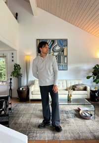 Man met tatoeages die een grijze lange mouwen shirt en zwarte broek draagt, staat in een moderne woonkamer met een witte bank en abstracte wandkunst.