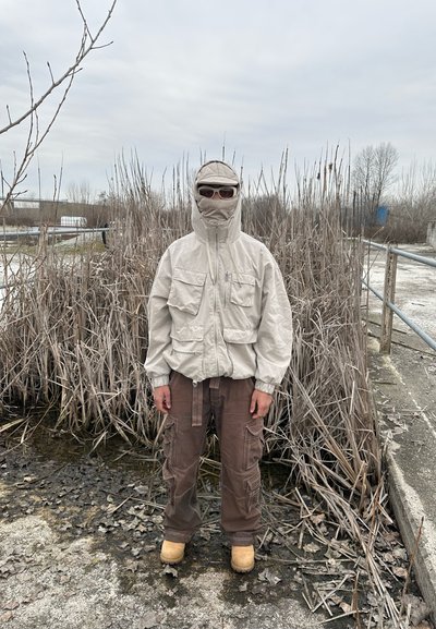 Persona con chaqueta beige, capucha, mascarilla y gafas de sol de pie sobre suelo embarrado con juncos secos en el fondo bajo un cielo nublado.