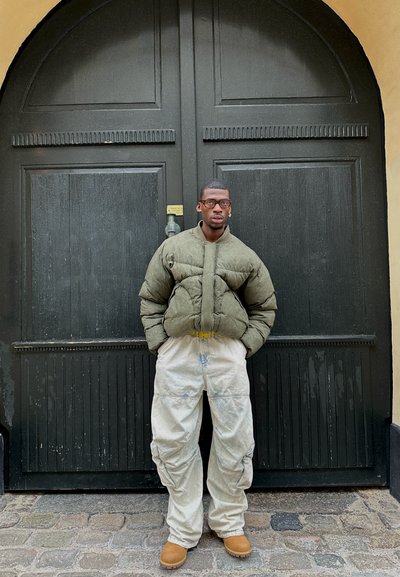 Chaqueta de plumas verde oliva con cuello alto, pantalones anchos blancos y botas de color canela, de pie frente a una gran puerta de madera negra.