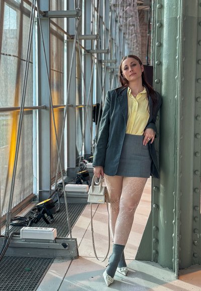 Mujer con blusa amarilla, blazer gris, minifalda, medias de encaje y tacones blancos apoyada en una columna metálica en un pasillo industrial de cristal.