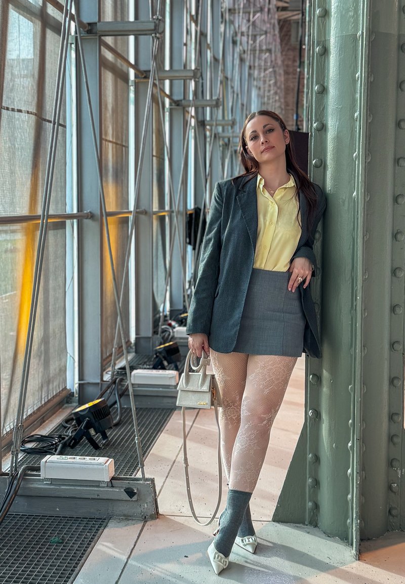 Femme en blouse jaune, blazer gris, mini-jupe, collants en dentelle et talons blancs s'appuie contre un pilier métallique dans un couloir industriel vitré.