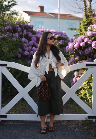 Mujer con gafas de sol y una blusa blanca anudada, de pie frente a una puerta blanca con arbustos florecidos morados detrás de ella.