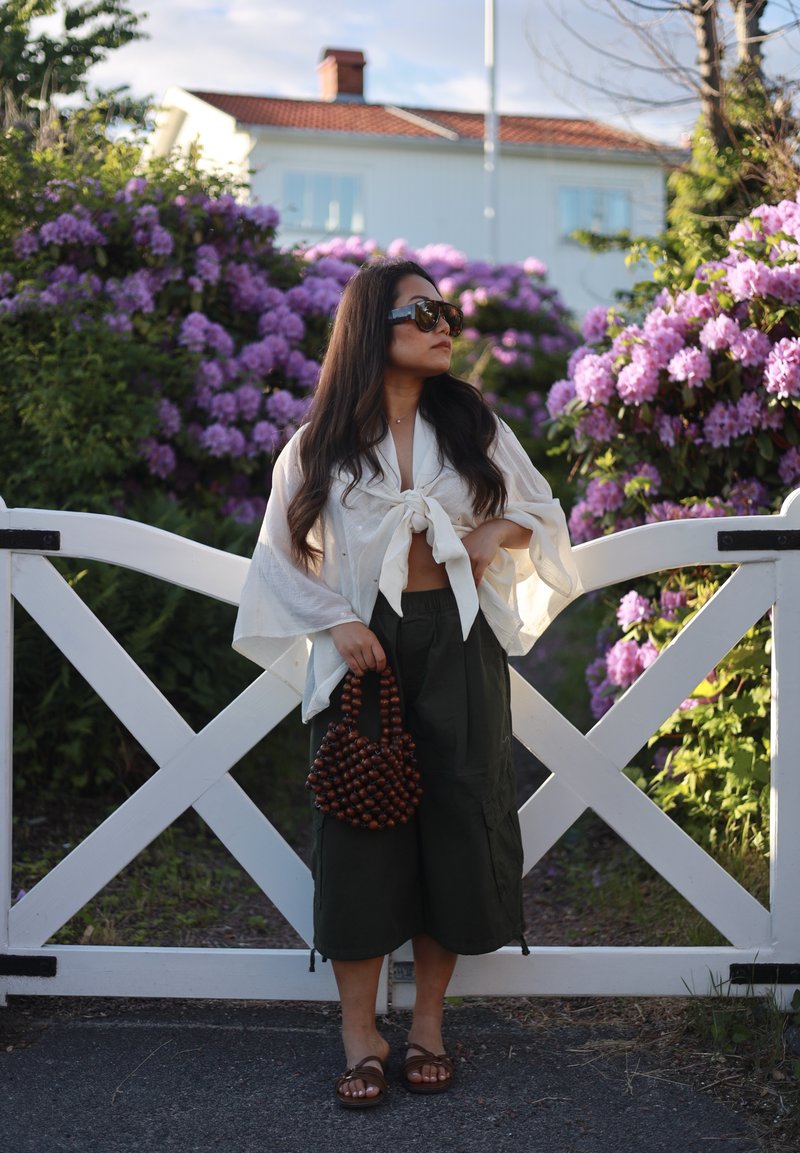Femme portant des lunettes de soleil et un chemisier blanc noué se tient devant une porte blanche avec des buissons fleurissant en violet derrière elle.