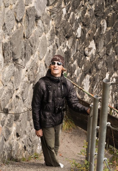 Persona con una chaqueta acolchada negra y pantalones verdes de pie en una escalera de piedra. Lleva un gorro oscuro y gafas de sol, sosteniendo una cámara.