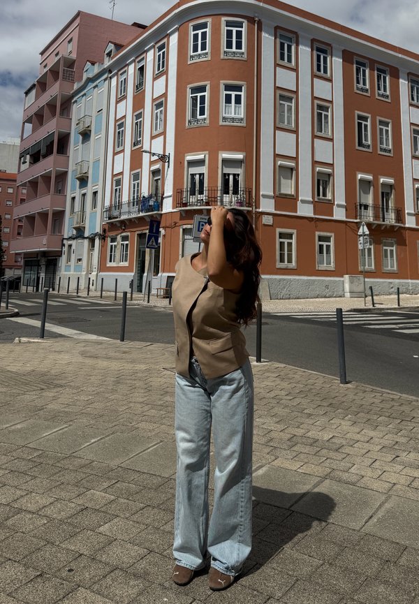 Femme aux longs cheveux foncés, vêtue d'un gilet beige et d'un jean large bleu clair, debout à un coin de rue pavé avec des immeubles à plusieurs étages en arrière-plan.