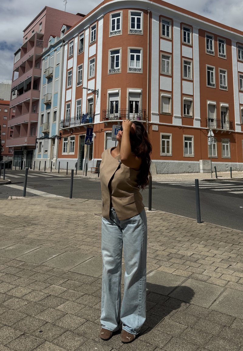 Woman with long dark hair in beige vest and wide light blue jeans standing on a paved street corner with multi-story buildings in the background.