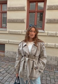 Mladá žena s vlnitými vlasmi, oblečená v nadmernej béžovej trenčkote a slnečných okuliaroch na hlave, stojaca na dlažobnej ulici pri béžovej budove.