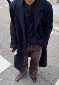 Dlhý čierny kabát, námornícky modrý top a hnedé manžestrové nohavice. Čierne topánky a slnečné okuliare. Drží biely pohár na kávu.