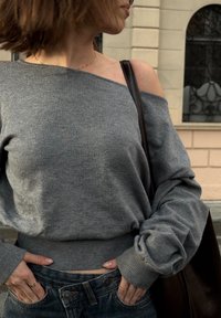 Donna che indossa un maglione grigio scivolato sulle spalle e jeans di denim con le mani nelle tasche, che porta una borsa in pelle marrone scuro.
