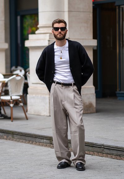 Hombre con barba y gafas de sol que lleva un cárdigan negro, camisa blanca, pantalones anchos beige y zapatos negros, de pie en la acera de la ciudad.