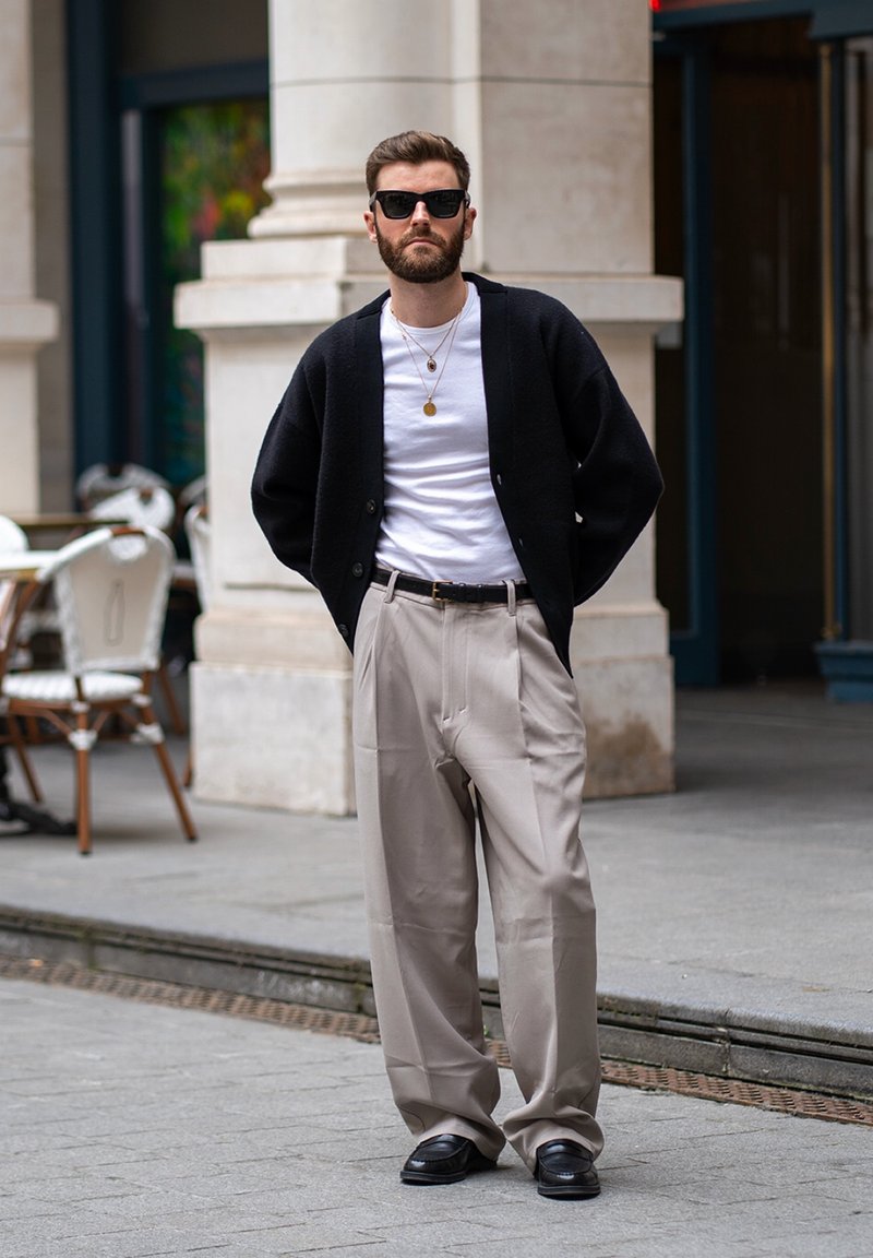 Homme avec barbe et lunettes de soleil portant un cardigan noir, une chemise blanche, un pantalon large beige et des chaussures noires, debout sur un trottoir en ville.