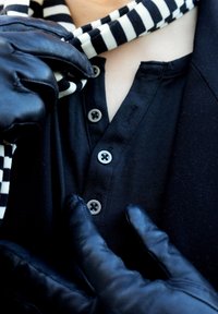 Chemise henley noire avec trois boutons gris, recouverte de gants en cuir noirs. Écharpe rayée noir et blanc drapée autour du cou.