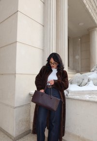 Femme en manteau de fourrure marron foncé et lunettes de soleil noires tenant un grand sac à main tressé marron, debout près de colonnes en pierre et d'une sculpture couverte de neige.