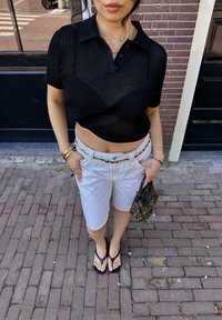 Zwarte mesh polo met een cropped fit, witte denim shorts, zwarte flip-flops en een gestreepte handtas. Achtergrond van bakstenen straat.