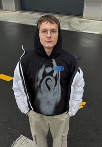 Zwarte hoodie met grafisch ontwerp en blauwe logo, gedragen onder een witte pufferjas, gecombineerd met beige cargobroek. Effen achtergrond.