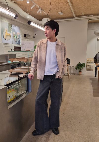 Joven con cárdigan beige y vaqueros negros anchos está junto al mostrador de la cafetería con bebidas embotelladas y menú en la pared.