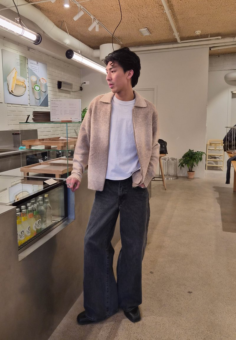 Jeune homme en cardigan beige et jeans noirs larges se tient au comptoir du café avec des boissons en bouteille et un menu au mur.