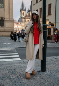 Beige παλτό trench πάνω από κόκκινο πουλόβερ, κρέμα παντελόνι σε φαρδιά γραμμή και καφέ μποτάκια μέχρι τον αστράγαλο. Ένα ελαφρύ καπέλο και τσάντα crossbody συμπληρώνουν την εμφάνιση.