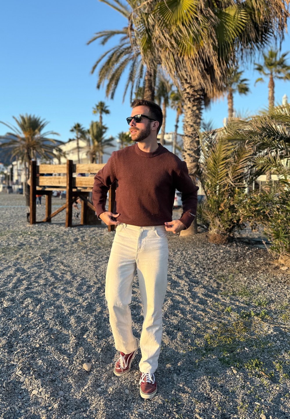 Homme portant des lunettes de soleil, un pull marron, un pantalon blanc et des baskets rouges, debout sur une plage rocheuse avec des palmiers en arrière-plan.