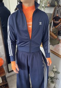 Tuta Adidas blu navy con strisce bianche, giacca con zip e pantaloni coordinati. Top sportivo arancione a strati sotto. Accento arancione brillante sul polso.