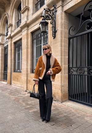 Brown shearling jacket with a soft interior, paired with black wide-leg jeans and a small black handbag, set against a backdrop of stone architecture.