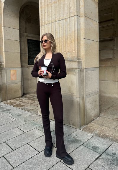 Conjunto de dos piezas de punto acanalado marrón oscuro con mangas largas, pantalones ajustados y zapatos de plataforma negros gruesos. Sosteniendo una taza de café.