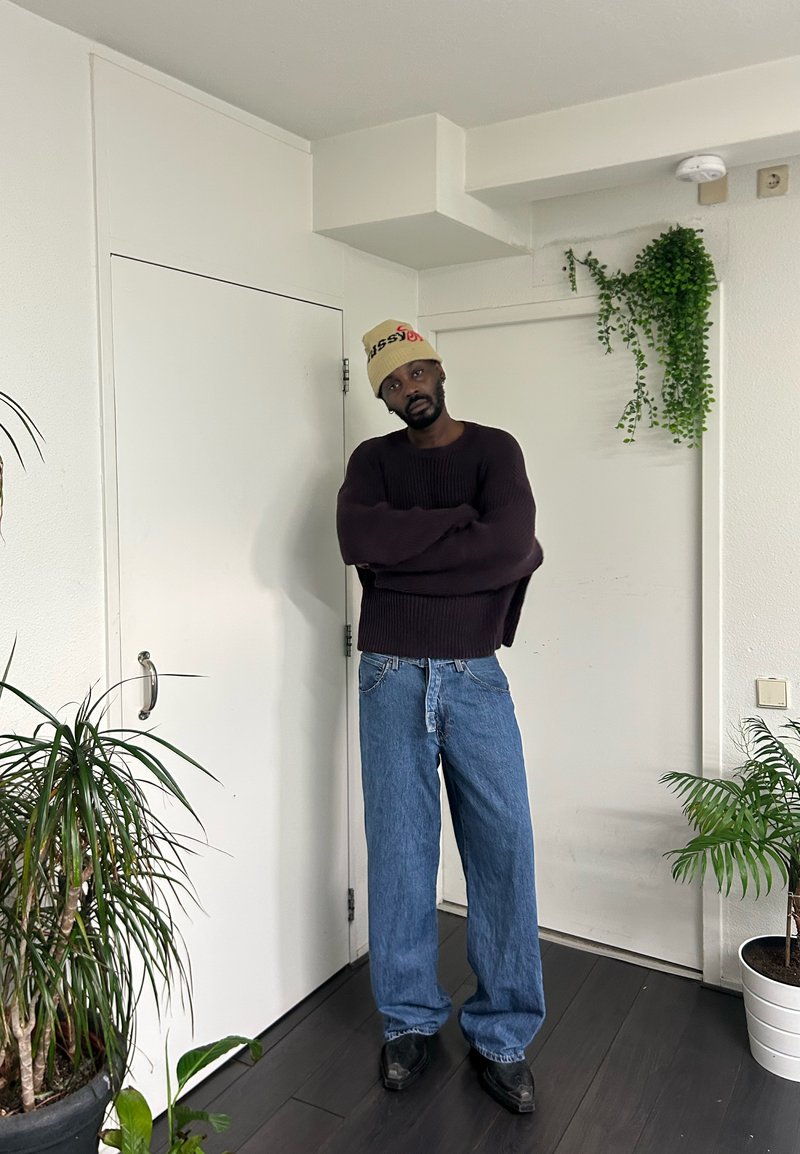 Dunkellila gerippter Pullover, hellblaue lockere Jeans und schwarze Schuhe. Das Model trägt eine beige Mütze. Innenraum mit Pflanzen und einer weißen Tür.